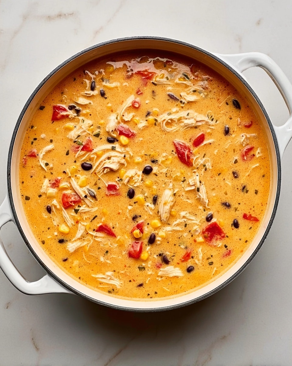 A white round pot filled with thick creamy soup showing several layers: a light orange creamy base with shredded white chicken pieces spread evenly, bright yellow corn kernels scattered throughout, whole black beans mixed in, and small red tomato chunks floating on top. The surface has a slightly glossy texture with a few green pepper slices visible inside. The background is a white marbled surface. photo taken with an iphone --ar 4:5 --v 7