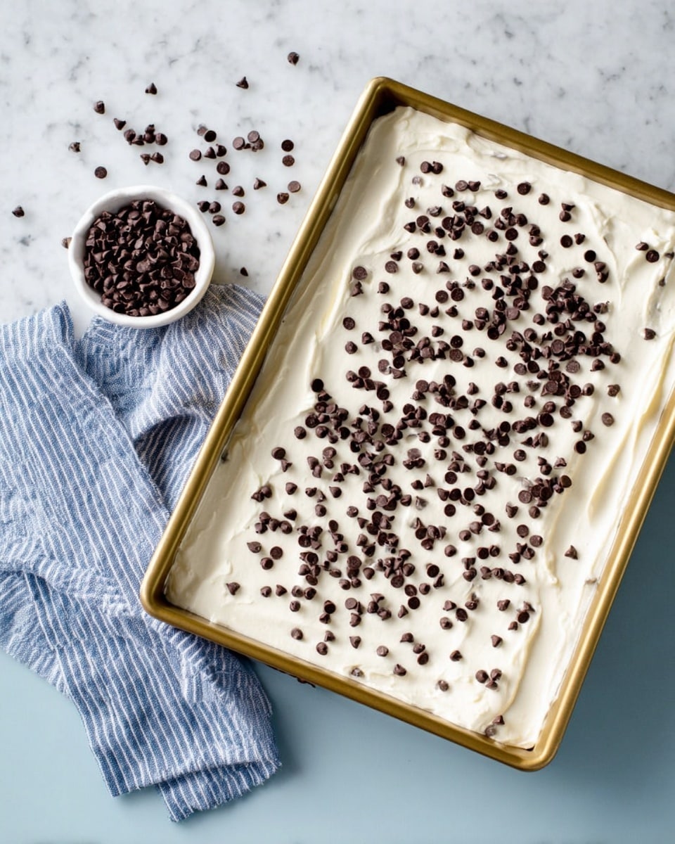 A rectangular dessert in a light gold baking tray with one thick layer of smooth white cream spread evenly on top, sprinkled with many small dark brown chocolate chips scattered randomly across the surface. Next to the tray is a small white bowl filled with the same dark chocolate chips, with some chips spilled around it. A blue and white striped cloth is placed to the side on a white marbled surface photo taken with an iphone --ar 4:5 --v 7