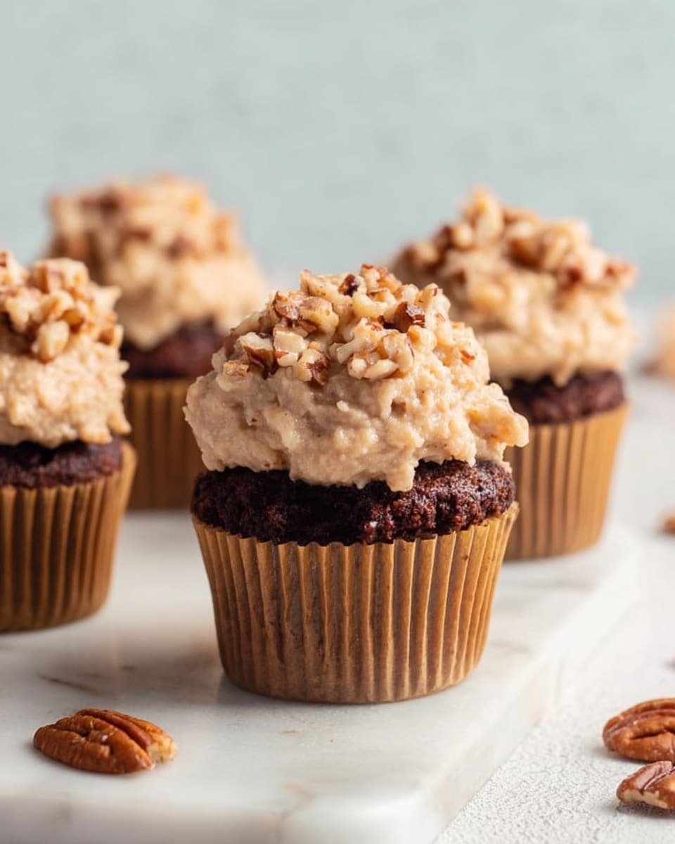 The image shows a close-up of four chocolate cupcakes arranged on a white marbled surface. Each cupcake has a golden brown paper liner. On top of each cupcake is a thick layer of beige frosting mixed with small pieces of chopped nuts, giving it a rough and chunky texture. The frosting is piled high, with the nuts creating small peaks and a slightly uneven surface. In the foreground, there are a few loose pecan pieces scattered around. The background is softly blurred, focusing attention on the cupcakes. Photo taken with an iphone --ar 4:5 --v 7