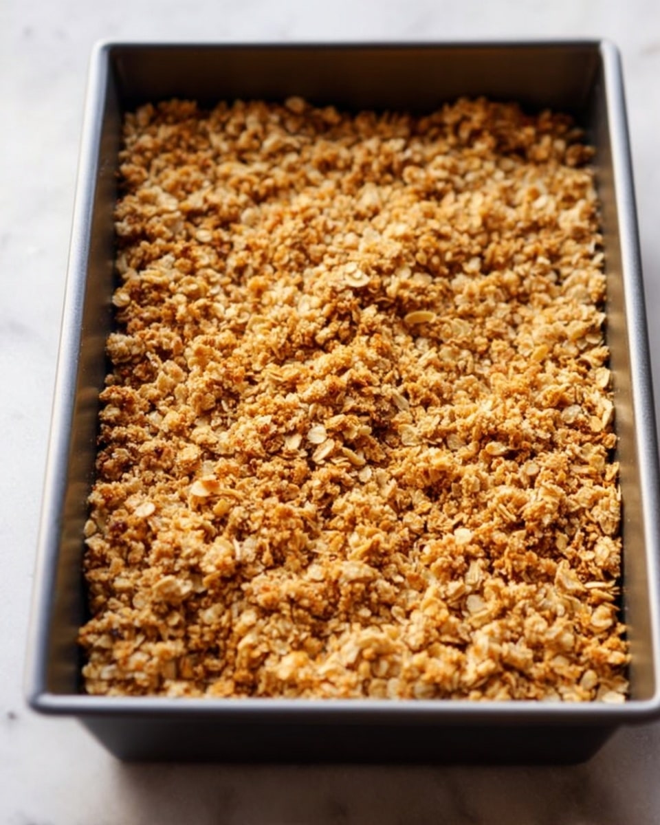 The image shows a rectangular metal baking pan filled with a crumbly oat topping that is golden brown and textured with small clusters, covering the entire surface evenly. The oat mixture has a rough, crunchy look with some toasted spots in a warm amber color, suggesting it is crispy. The pan is placed on a white marbled surface, and soft natural light highlights the crispy texture of the topping. Photo taken with an iphone --ar 4:5 --v 7