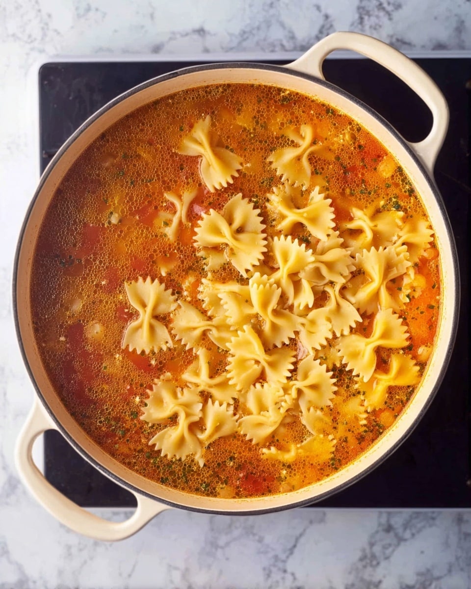 A round white pot with two handles is filled with a tomato-based broth that has a mix of orange and red hues with visible herbs and spices. On one side, uncooked farfalle pasta floats on the surface, showing its pale yellow color and distinct bow-tie shapes with ruffled edges. The pot sits on a black stove against a white marbled background. The stew’s surface shows some oil floating, giving a slight shine and texture to the broth photo taken with an iphone --ar 4:5 --v 7