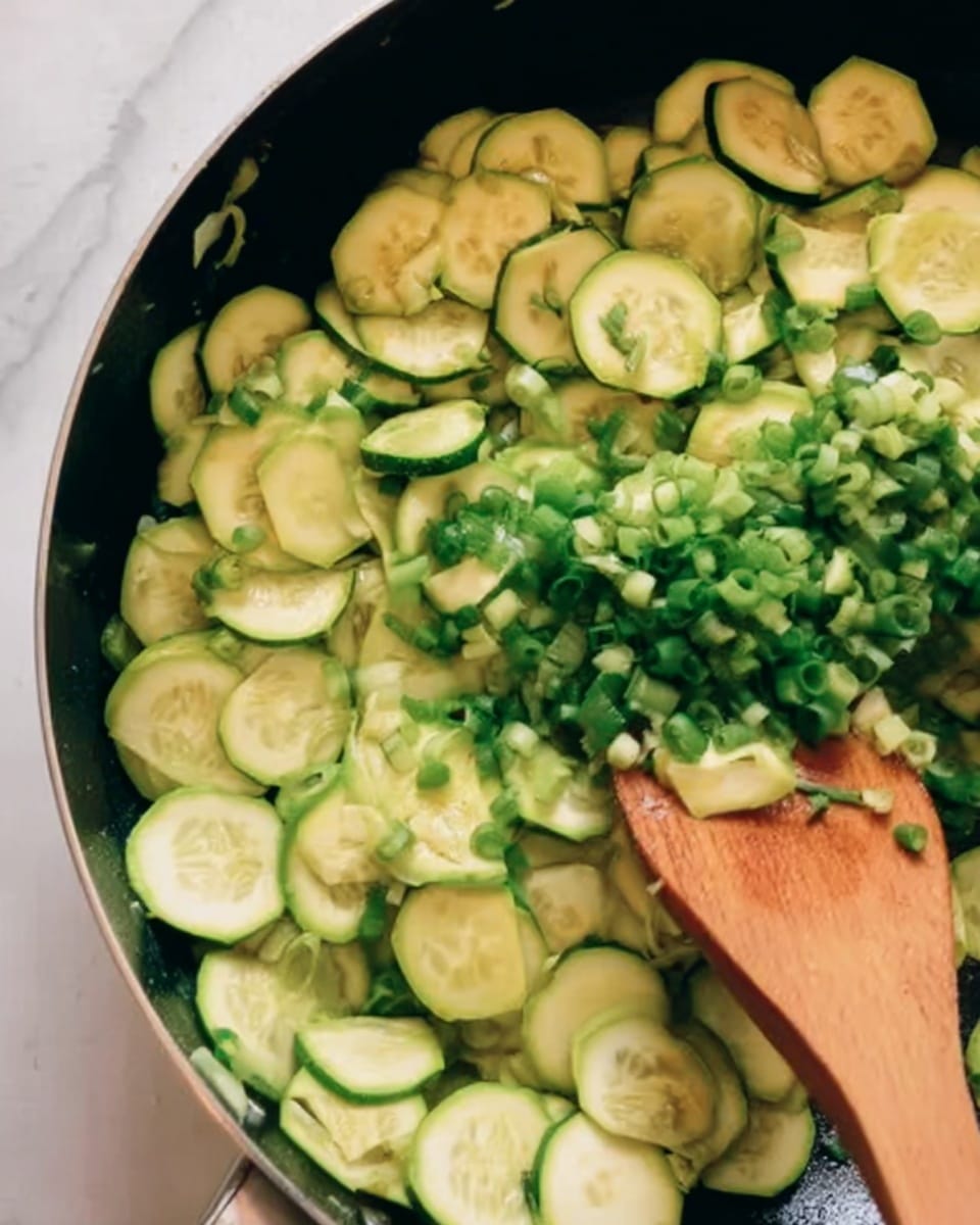 The image shows a close-up of a frying pan with thinly sliced green zucchini pieces spread evenly across the bottom layer. On top of the zucchini, there are finely chopped green onions being added by a wooden spatula held by a woman's hand coming from the bottom of the image. The zucchinis are light green with darker green edges and look slightly soft, while the chopped green onions add flecks of bright, fresh green color. The frying pan sits on a white marbled surface. Photo taken with an iphone --ar 4:5 --v 7