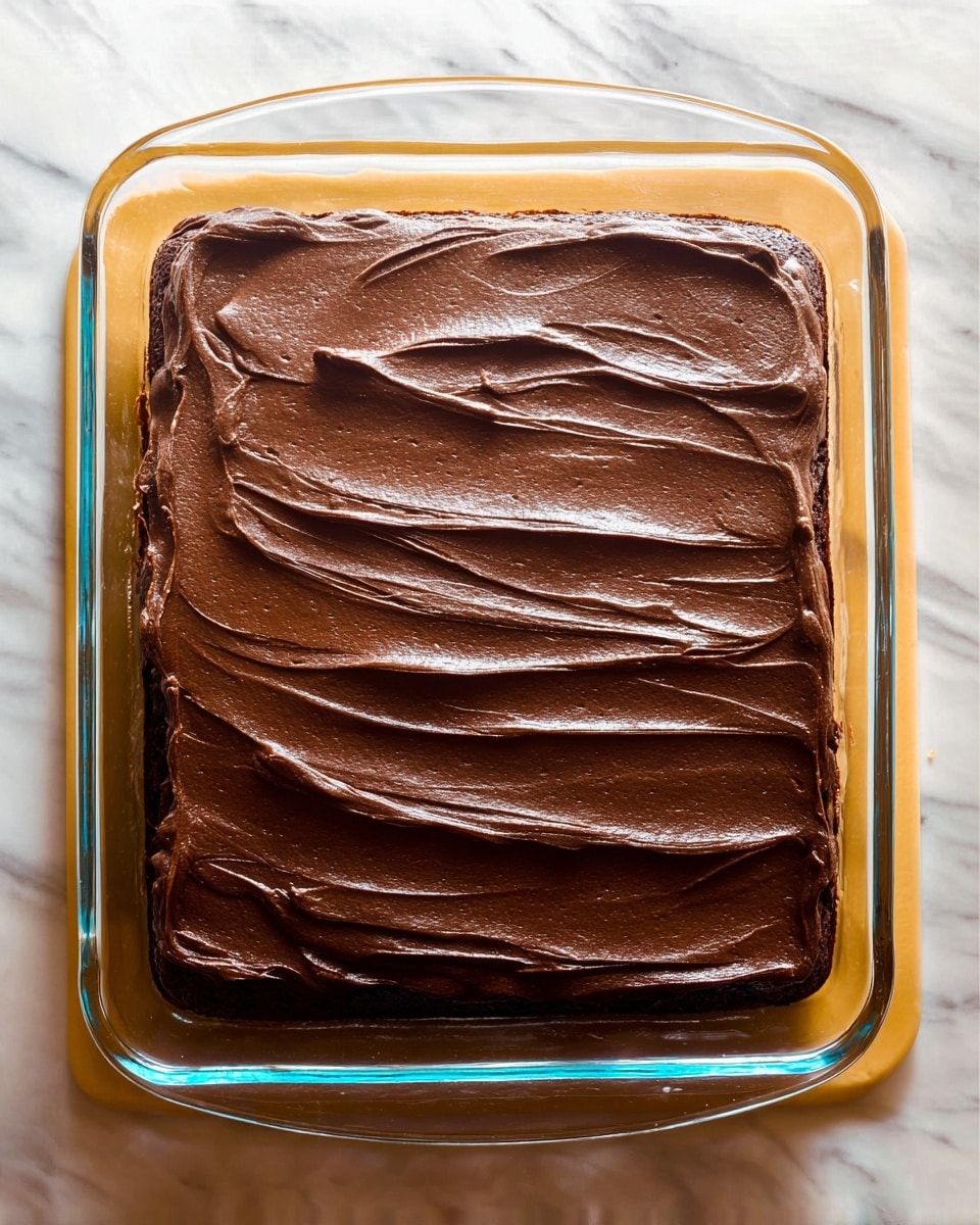 A clear glass rectangular dish holds a single-layer chocolate cake, covered smoothly with thick, dark brown chocolate frosting. The frosting is spread evenly with visible sweeping patterns on the top surface. The dish sits on a white marbled surface. photo taken with an iphone --ar 4:5 --v 7