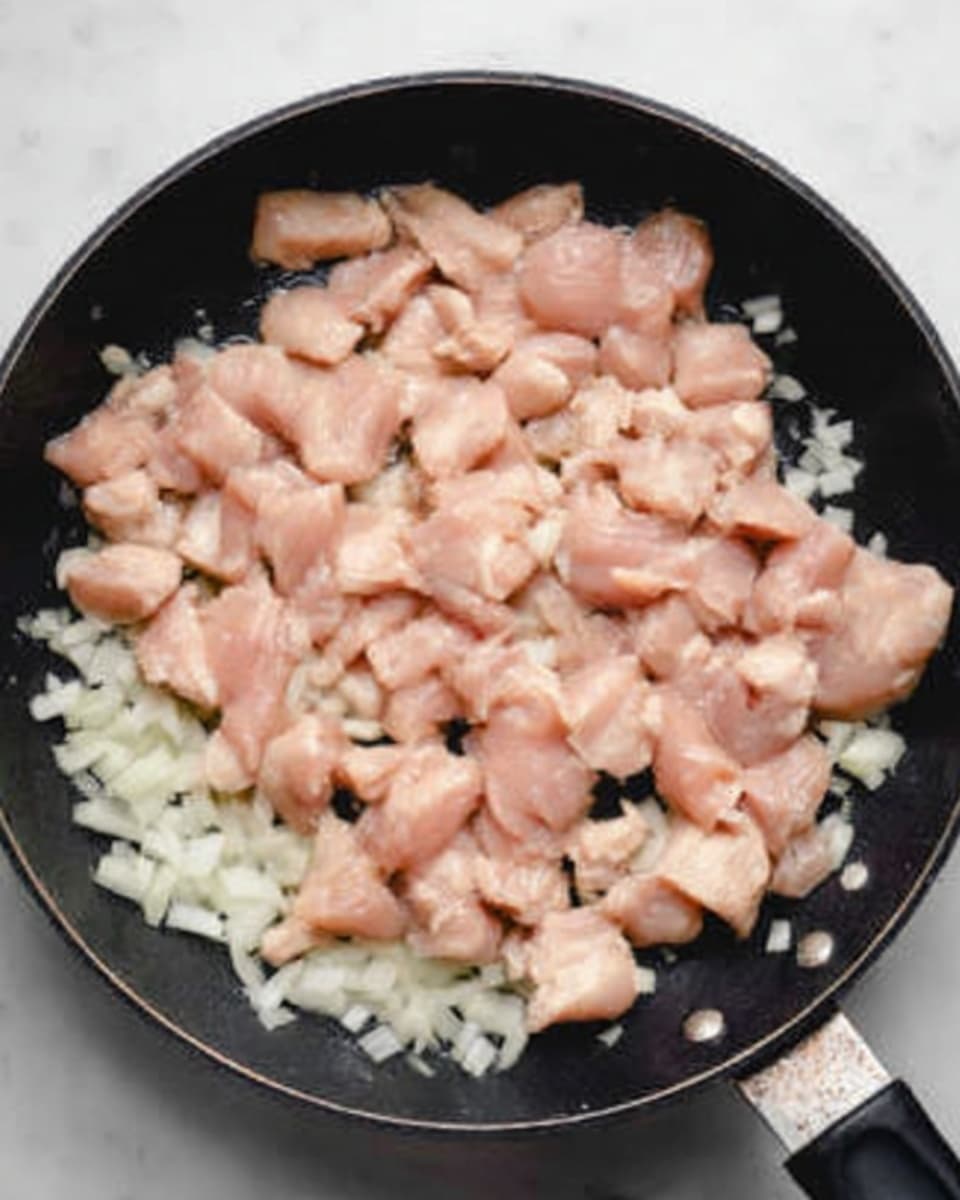 The image shows raw pieces of light pink chicken spread out over a layer of chopped white onions evenly placed at the bottom of a black frying pan. The chicken pieces vary in size and are spread across the entire pan, creating a contrast between the pale chicken and the white onions beneath. The frying pan has a smooth black texture and it sits on a white marbled surface. There is a glimpse of a woman's hand holding onto the frying pan's handle on the right side of the image. Photo taken with an iphone --ar 4:5 --v 7