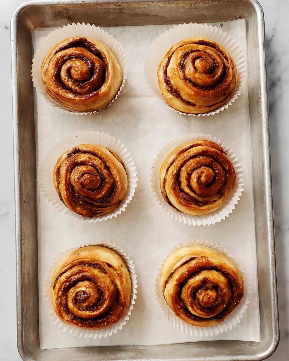 Six cinnamon rolls with a golden brown color and dark brown spiral lines are placed individually in white paper liners on a metal baking tray lined with white parchment paper. The rolls are evenly spaced in two rows of three on the tray, each showing a soft, slightly shiny surface with a swirled pattern. The background is a white marbled texture photo taken with an iphone --ar 4:5 --v 7