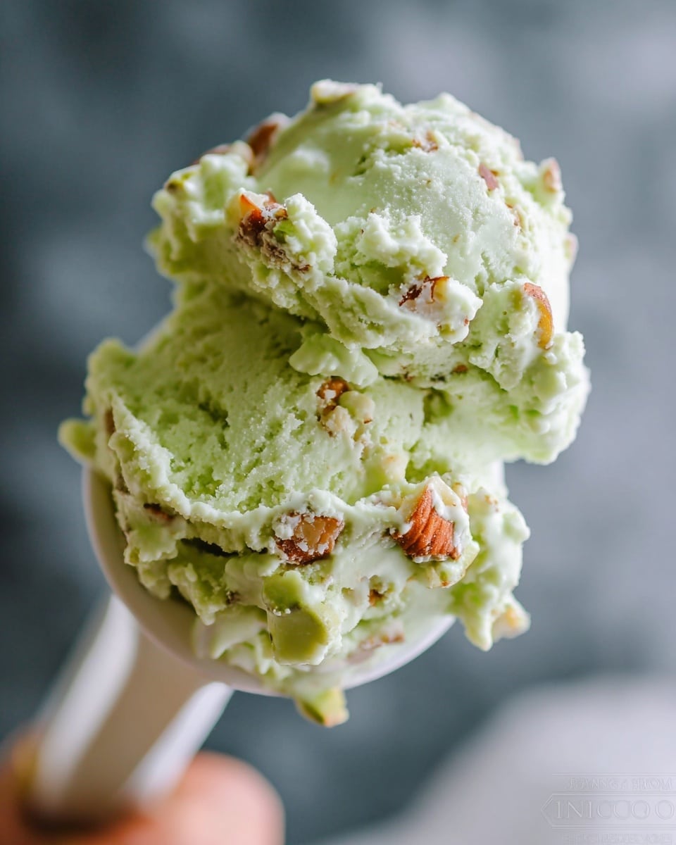 A close-up of a scoop of light green ice cream held by a woman's hand with a white spoon. The ice cream has a crumbly texture mixed with pieces of nuts and small white chunks, showing bits of orange and green colors embedded in it. The background features a soft blurred contrasting darker area with a white marbled surface below. The focus is sharp on the ice cream and spoon, highlighting the creamy and nutty details. photo taken with an iphone --ar 4:5 --v 7
