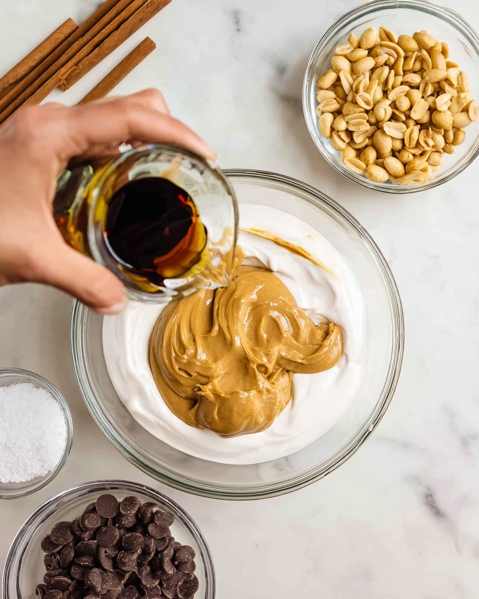 A clear glass bowl sits on a white marbled surface, holding several thick layers: a smooth white creamy base, a large dollop of thick beige peanut butter in the center, and a light brown syrup pooling around the edges. A woman's hand pours dark brown vanilla extract from a small glass container onto the peanut butter layer. Nearby, three smaller clear glass bowls contain different ingredients: one with roasted peanuts, one with dark chocolate chips, and one with white granulated sugar. Wooden sticks lie on the marbled surface in the top left corner. Photo taken with an iphone --ar 4:5 --v 7