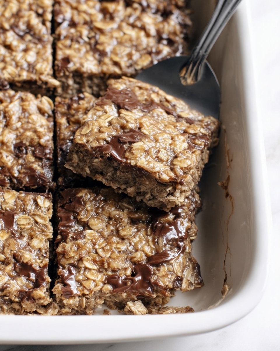 The image shows a close-up of a white baking dish with four thick oatmeal bars inside. Each bar has a rough texture with visible oats and a light brown color mixed with a darker, chocolatey layer. The bars are soft and slightly gooey, with one bar being lifted by a metal spatula, showing a chewy inside. The surface of the bars looks slightly shiny and moist, with some melted chocolate filling spots. The background is a smooth white marbled surface. photo taken with an iphone --ar 4:5 --v 7