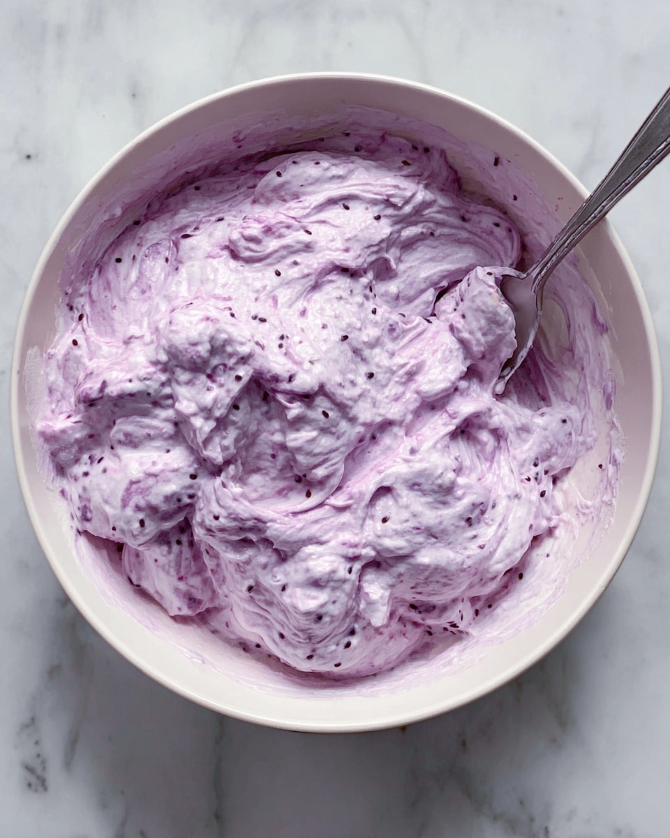 A round white bowl filled with a thick, creamy mixture that is light purple with streaks of darker purple swirled throughout. The mixture looks fluffy and textured, dotted with small seeds scattered inside. A silver spoon rests on the right side of the bowl, partially dipped in the mixture. The bowl is set on a white marbled surface. photo taken with an iphone --ar 4:5 --v 7