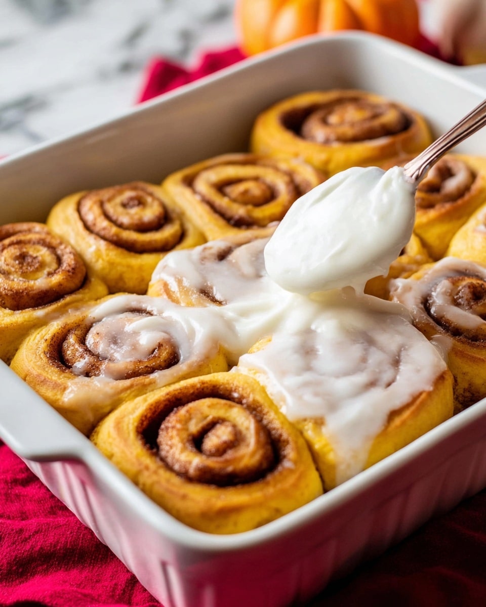 A white rectangular baking dish holds nine pumpkin cinnamon rolls arranged in three rows of three. Each roll has a golden orange base with a darker brown cinnamon spiral inside, showing a soft texture. A thick layer of glossy white icing is being spread on the top right rolls using a spoon, creating a smooth, creamy look. The dish sits on a bright red cloth, with the background showing a blurred white marbled texture. Photo taken with an iphone --ar 4:5 --v 7