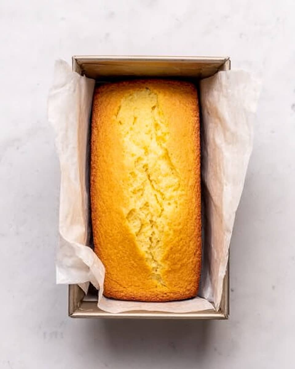 The image shows a golden yellow loaf cake with a slightly cracked top, sitting inside a rectangular metal baking pan lined with white parchment paper that extends over the edges. The pan is placed on a white marbled surface, and the cake has a smooth, soft texture with a light crust forming around the edges. The corners of the cake touch the pan closely, and the overall appearance is fresh and warm. Photo taken with an iphone --ar 4:5 --v 7