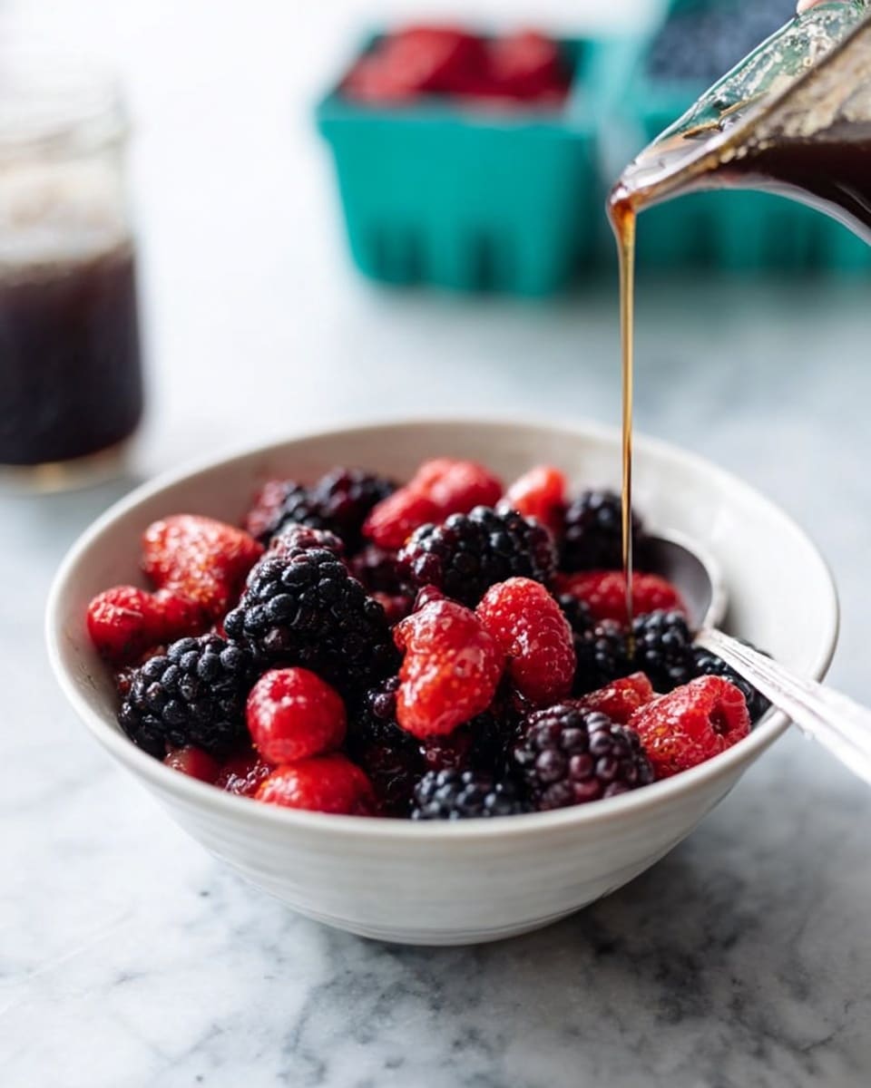 A white bowl filled with fresh mixed berries including blackberries and raspberries, sitting on a white marbled surface. The berries have a rich, dark purple and bright red color, and the texture looks soft and juicy. A silver spoon rests inside the bowl on the right side. A woman's hand is pouring a dark syrup over the berries from above, with a small glass jar of syrup blurred in the background. Some blurred berry containers in teal are also seen in the background, creating a light and fresh setting. Photo taken with an iphone --ar 4:5 --v 7