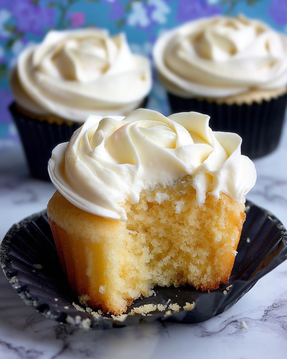 The image shows a close-up of a cupcake with two layers: the bottom layer is a light yellow cake with a soft texture, and the top layer is a thick swirl of creamy white frosting shaped like a rose, with smooth and slightly shiny petals. One cupcake has a bite taken from it, revealing the moist inside of the cake. The cupcakes rest in black paper liners, placed on a white marbled surface with a blue and purple floral pattern in the background. Photo taken with an iphone --ar 4:5 --v 7