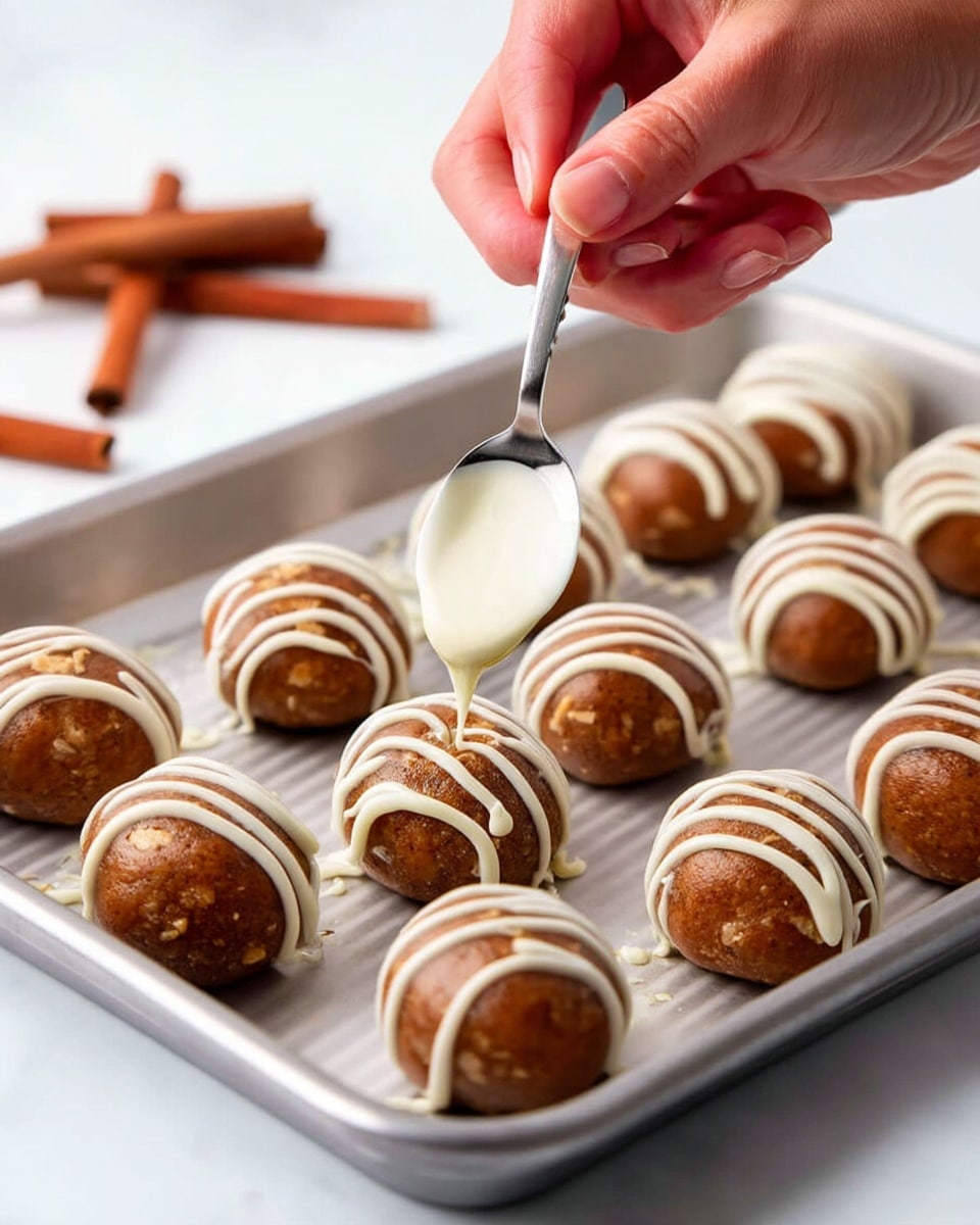 The image shows a metal tray with two rows of light brown round balls that have a rough texture with small pieces visible inside. The balls are being decorated with white creamy drizzle in curved lines. A spoon is held above one ball by a woman's hand, pouring the white drizzle over it. The background is a white marbled surface with two cinnamon sticks blurred in the upper left corner. The scene is bright and clear, focusing closely on the balls and the spoon. photo taken with an iphone --ar 4:5 --v 7