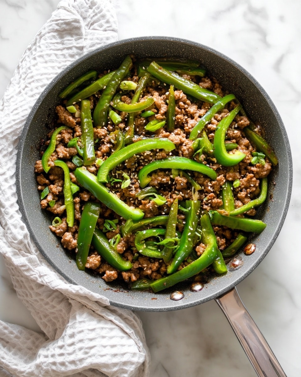 The image shows a gray pan filled with cooked ground meat mixed with long slices of green bell pepper spread evenly throughout. The meat is browned and crumbled into small pieces, while the green peppers add a fresh contrast. On top, there are small sprinkles of sesame seeds and finely chopped green onions scattered across the dish. A white cloth with a textured pattern is partially wrapped around the pan's handle, resting on a white marbled surface. photo taken with an iphone --ar 4:5 --v 7