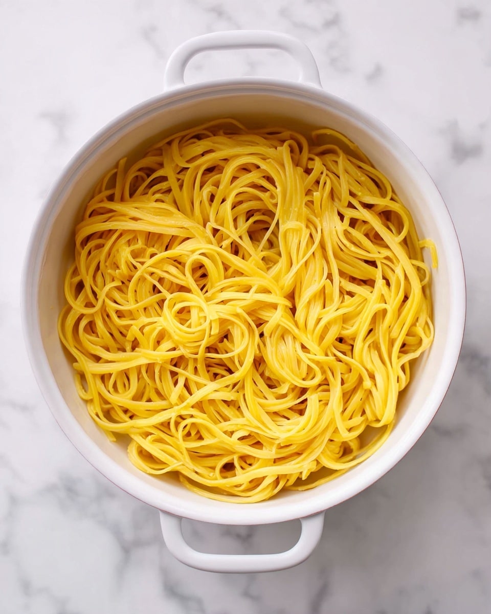 A white pot filled with a single layer of cooked yellow pasta noodles that look soft and slightly shiny, sitting on a white marbled surface. The noodles are long, thin, and slightly twisted with some overlapping, creating a simple but inviting look. Photo taken with an iphone --ar 4:5 --v 7