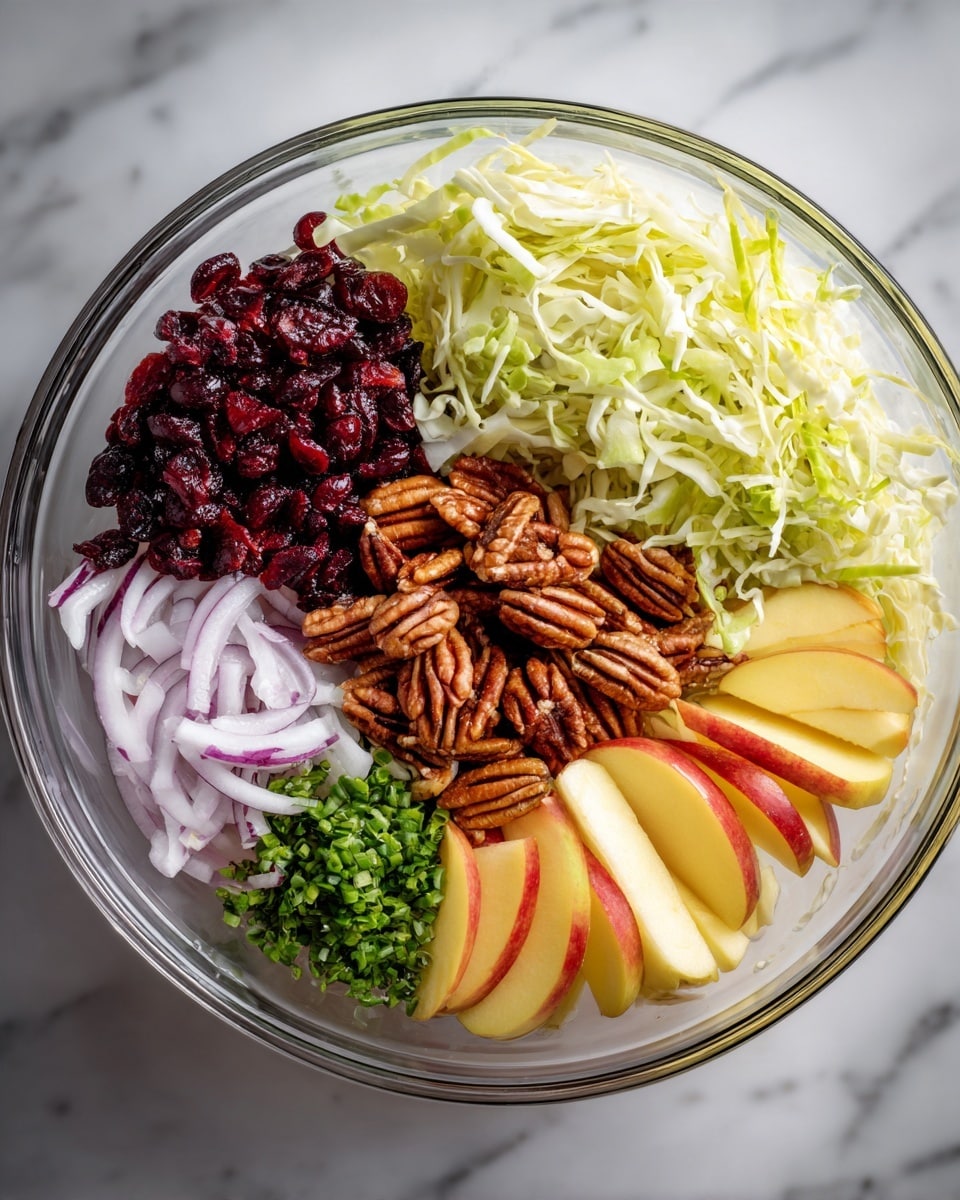 The image shows a clear glass bowl filled with seven different ingredients neatly placed in sections, viewed from above on a white marbled surface. There is a large pile of shredded light green cabbage covering nearly a third of the bowl on the top right. To the left of the cabbage, dark red dried cranberries form a small pile. Below the cranberries are thin slices of pale purple onion. Next to the onions on the bottom left, there is a small bunch of bright green chopped herbs. The center of the bowl holds a heap of toasted brown pecans. On the bottom right, two types of apple slices are placed: yellowish slices with some red skin on the edge and pale yellow slices without skin. photo taken with an iphone --ar 4:5 --v 7
