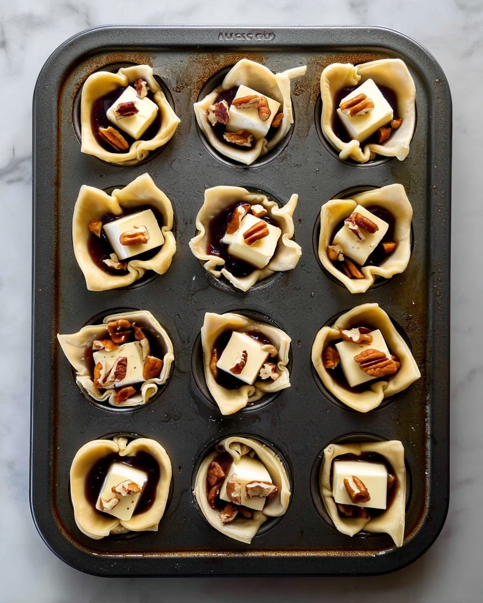 The image shows a dark metal muffin tray holding twelve small pastry cups. Each pastry cup is pale beige with a slightly crumpled texture, forming a flower-like shape around the edges. Inside each cup, there is a glossy dark brown layer of syrupy sauce at the bottom. On top of this, a small square of white, smooth butter is placed in the center. Around and on top of the butter, there are small pieces of chopped pecans scattered irregularly, adding a rough texture and warm brown color contrast. The tray sits on a white marbled surface. Photo taken with an iphone --ar 4:5 --v 7