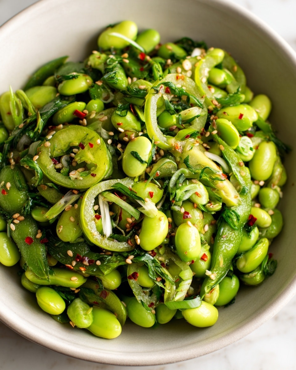 A close-up view of a fresh green salad inside a white bowl placed on a white marbled surface. The salad is made of bright green edamame beans as the base layer mixed with thinly sliced green peppers and scallions arranged on top. The layers are tossed with small green herbs and lightly dressed with hints of chili flakes and sesame seeds scattered throughout, creating pops of white and red colors. The salad has a shiny, slightly oily texture that highlights the freshness of the greens. photo taken with an iphone --ar 4:5 --v 7