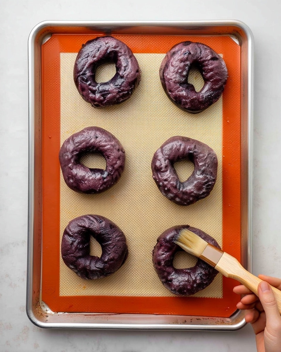 On a silver baking tray lined with a tan silicone mat that has an orange border, there are six raw dark purple dough rings, arranged in two rows of three. The dough rings have a slightly rough texture with small darker spots. At the bottom right corner, a woman's hand holds a wooden brush applying liquid to one of the dough rings. The tray rests on a white marbled surface. photo taken with an iphone --ar 4:5 --v 7
