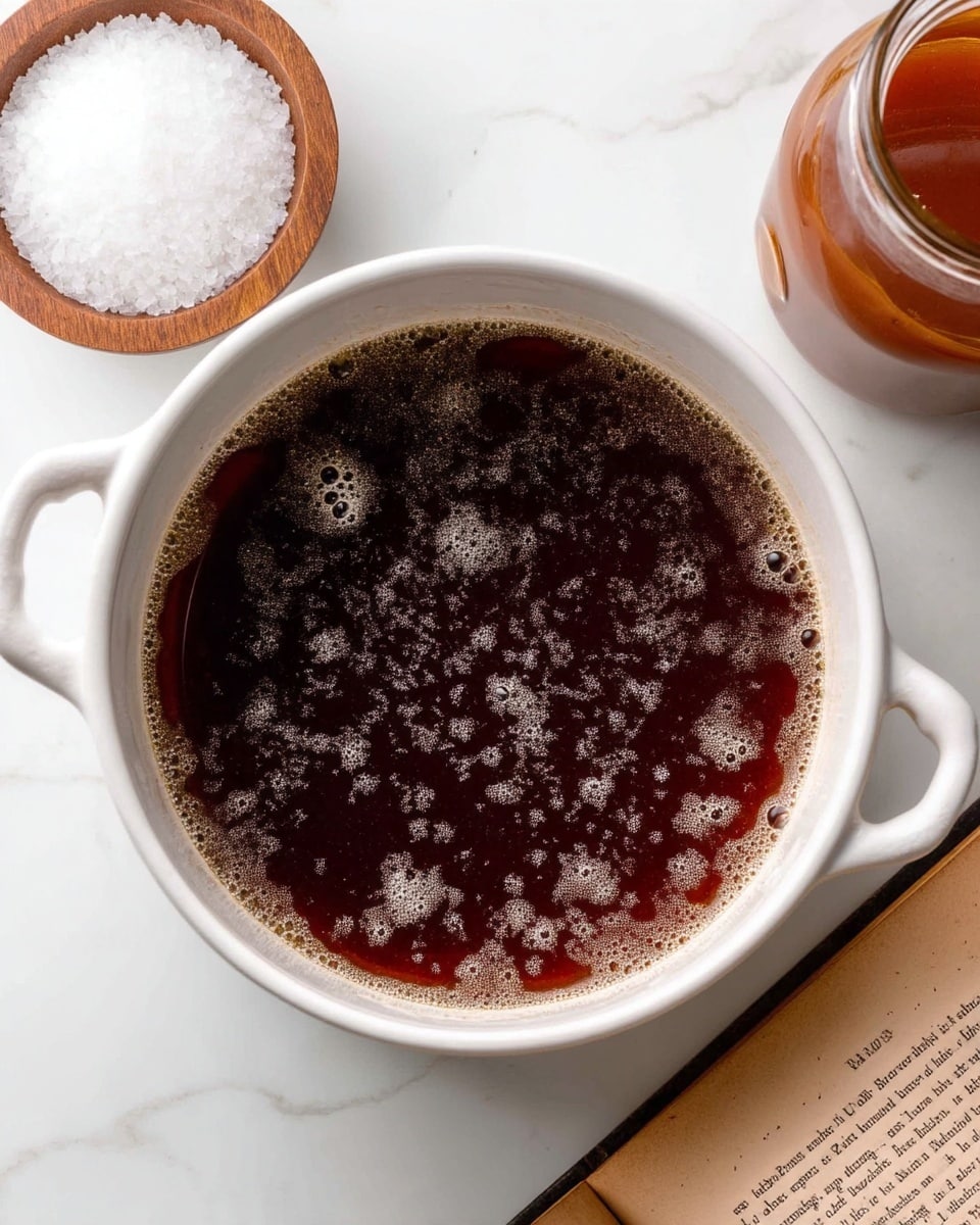 A white pot filled with dark brown liquid that has light bubbles floating on top, creating a slightly foamy texture. The pot sits on a white marbled surface, surrounded by a small wooden bowl filled with coarse white salt on the bottom left and an old open book page on the bottom right. A jar filled with a reddish-brown sauce or liquid is partly visible in the top right corner. Photo taken with an iphone --ar 4:5 --v 7