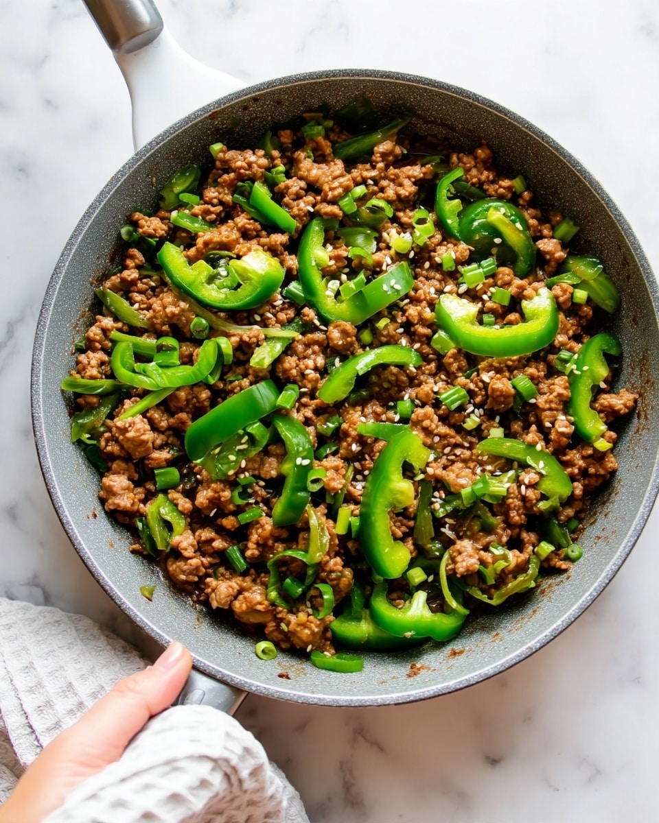 The image shows a gray frying pan filled with cooked ground meat that is brown and crumbly in texture. Mixed within the meat are slices of bright green bell peppers, some cut into strips and others into small pieces, adding patches of green color throughout. The dish is sprinkled with small bits of light green chopped scallions and a few white sesame seeds, creating small contrasting highlights. The pan sits on a white marbled surface, and a woman's hand holding a white textured cloth is visible gripping the pan handle on the lower left side of the image. photo taken with an iphone --ar 4:5 --v 7