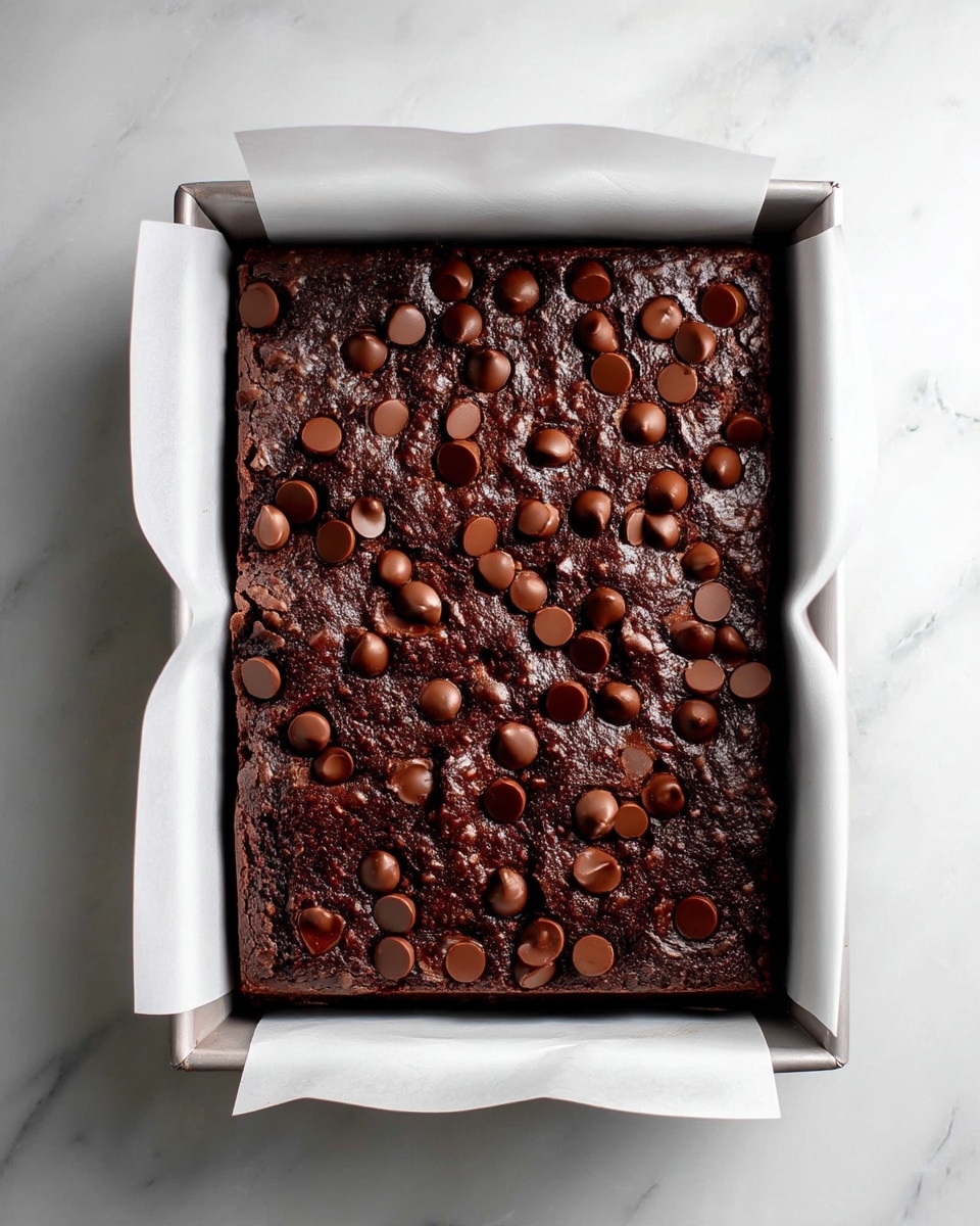A rectangular baking pan lined with white baking paper holds a freshly baked dark chocolate brownie. The brownie has a rough, textured top layer with small, round chocolate chips scattered across the surface. The dark brown color of the brownie contrasts with the white baking paper, which folds over the edges of the pan. The pan is placed on a white marbled surface, and the overall look is clean and simple. Photo taken with an iphone --ar 4:5 --v 7