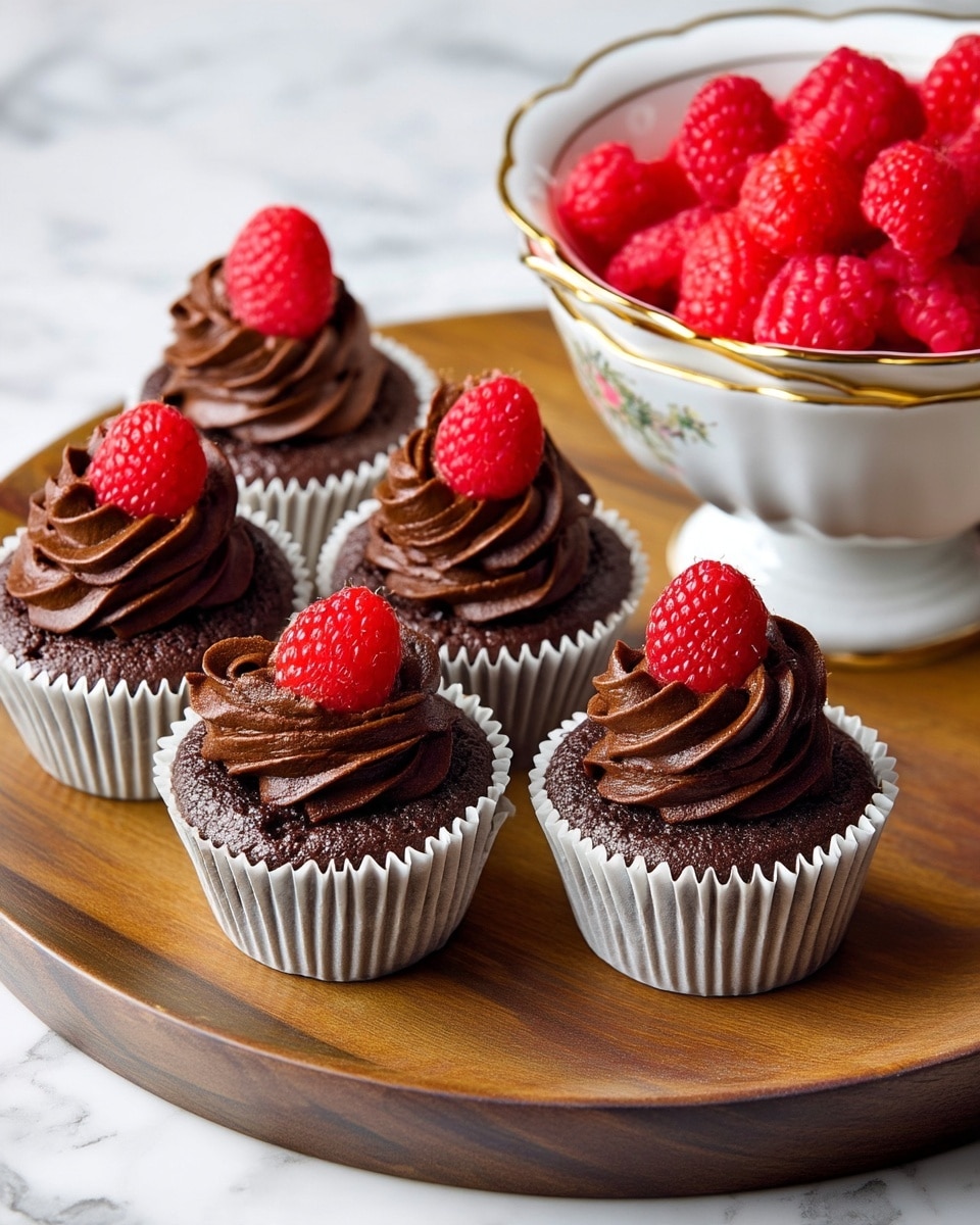The image shows five small chocolate cupcakes arranged on a round wooden tray. Each cupcake has a single layer of dark chocolate cake topped with a swirl of smooth, rich chocolate frosting. On top of each frosting swirl sits a bright red raspberry, adding a fresh pop of color. The cupcakes are in white paper liners with slightly ruffled edges. To the right, a white bowl with gold trim is filled with more fresh raspberries. The background is a white marbled surface. photo taken with an iphone --ar 4:5 --v 7