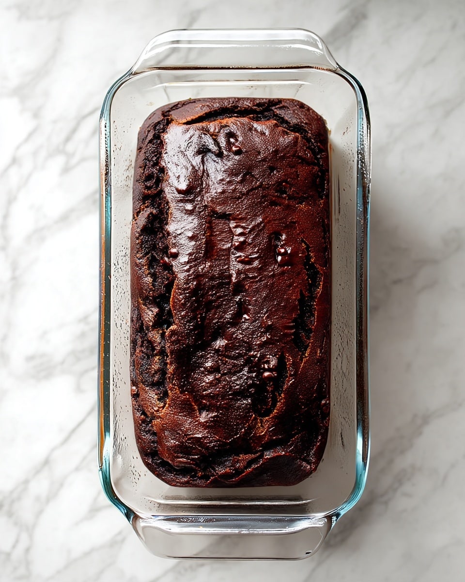 A single dark brown chocolate loaf cake sits in a clear glass baking dish. The cake has a rough, slightly cracked top with a dense texture and small uneven bumps across the surface. The glass dish has handles on both ends and is on a white marbled surface. The lighting shows the rich, deep color of the cake clearly. photo taken with an iphone --ar 4:5 --v 7