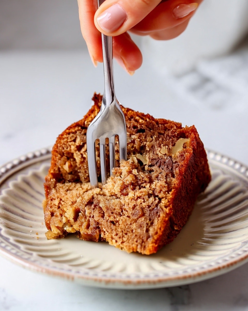 A close-up image of a single slice of brown cake with a moist and textured inside, showing small bits of nuts, placed on a white plate with a ridged edge. A silver fork is cutting into the cake from the right side, held by a woman's hand. The background is softly blurred with a white marbled texture underneath the plate. Photo taken with an iphone --ar 4:5 --v 7