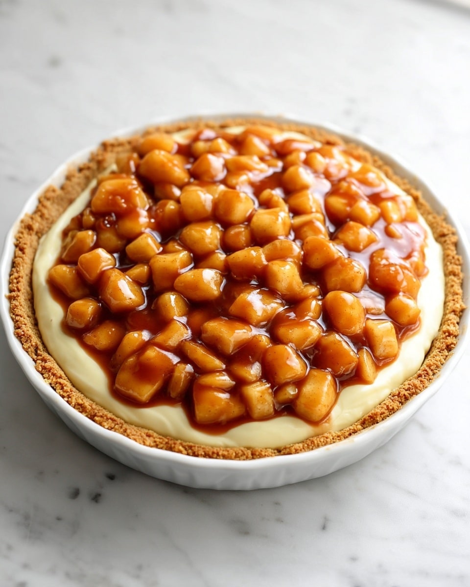 A white ceramic pie dish holds a three-layer dessert on a white marbled surface. The bottom layer is a crumbly light brown pie crust. The middle layer is a smooth, pale yellow cream filling, evenly spread over the crust. The top layer is a generous amount of small apple chunks coated in a shiny caramel sauce, covering the cream fully but not overflowing the crust edges. The pie looks fresh and inviting. Photo taken with an iphone --ar 4:5 --v 7
