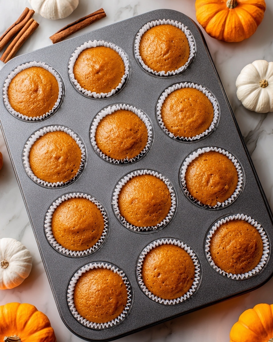 A dark grey metal muffin tray filled with 12 white paper liners, each holding smooth, glossy orange batter that is neatly rounded on top, with slight uneven textures on some surfaces. The tray sits on a white marbled surface. Around the edges, there are small white and orange pumpkins and two cinnamon sticks in the upper left corner, adding a cozy autumn feel to the scene. photo taken with an iphone --ar 4:5 --v 7