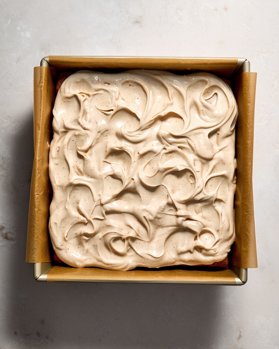 A close-up view of a single layer of creamy light brown batter with small darker specks spread thickly in a rectangular baking pan lined with brown parchment paper, the surface of the batter showing soft, swirling patterns and peaks. The pan sits on a white marbled textured surface. photo taken with an iphone --ar 4:5 --v 7