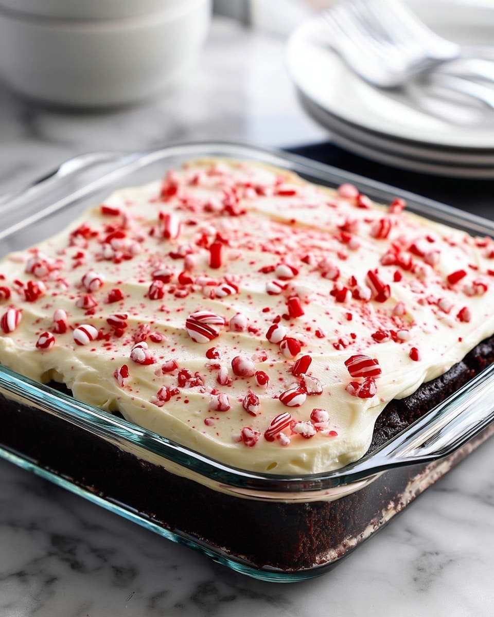 A square glass baking dish filled with two layers: the bottom layer is dark, rich chocolate cake with a soft texture, and the top layer is thick, creamy white frosting spread evenly. The frosting is topped with small, bright red and white crushed candy pieces scattered all over. The dish is placed on a white marbled surface, with blurred white plates and silverware in the background. Photo taken with an iphone --ar 4:5 --v 7