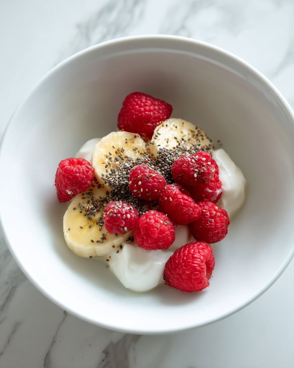 A white bowl holds a simple dish with three main layers. At the bottom, there are white yogurt dollops. On top of the yogurt is a half banana, pale yellow with a soft texture. The top layer is a mix of bright red raspberries and small black chia seeds scattered over the banana and yogurt. The bowl sits on a white marbled surface. photo taken with an iphone --ar 4:5 --v 7