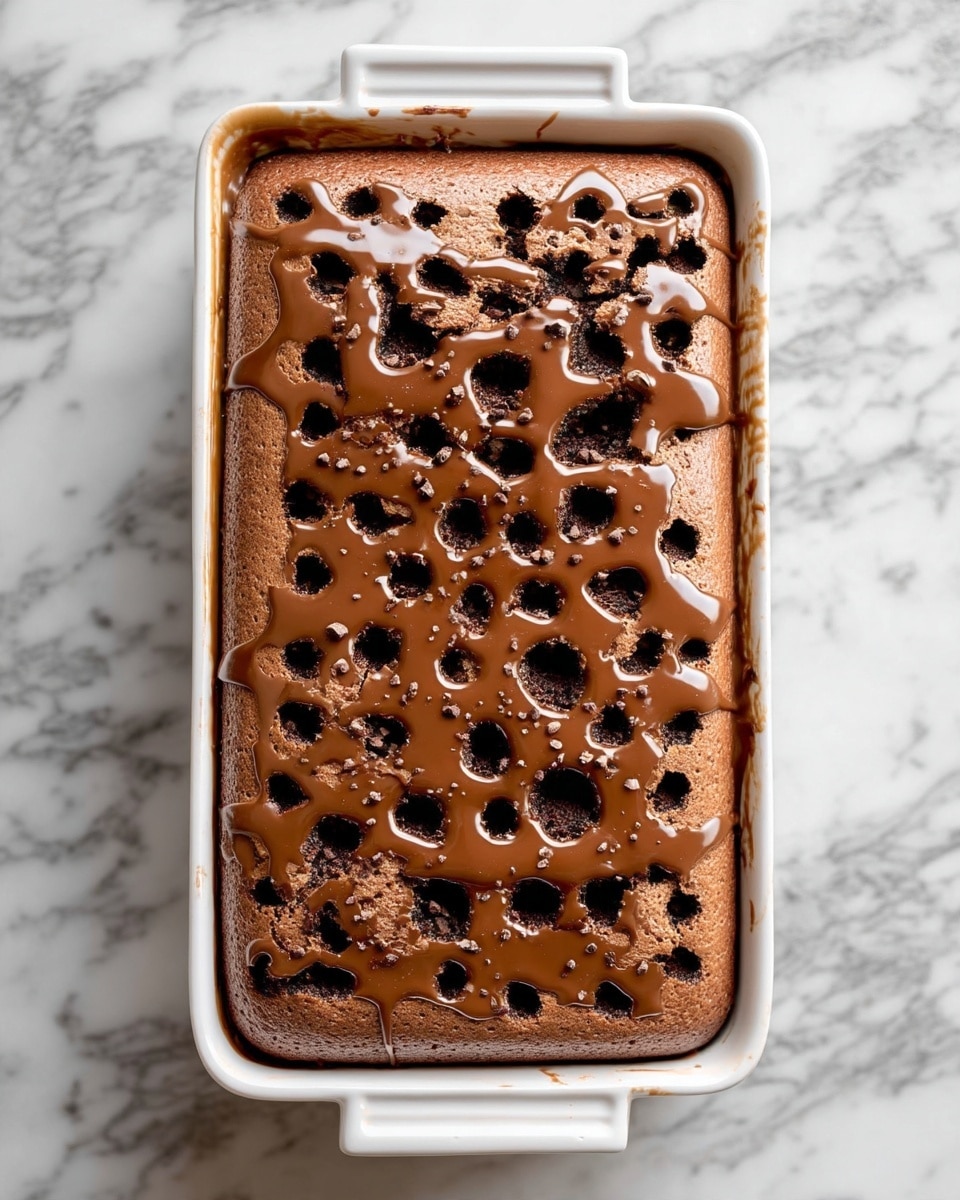 The image shows a large rectangular chocolate dessert in a white baking dish. It has one main layer of soft, light brown chocolate cake with many evenly spaced holes poked on top. Inside these holes, dark chocolate or syrup fills them, adding a glossy dark contrast to the matte surface. The whole top is covered with a shiny light brown chocolate glaze that drips slightly on the edges. The background is a white marbled surface. Photo taken with an iphone --ar 4:5 --v 7