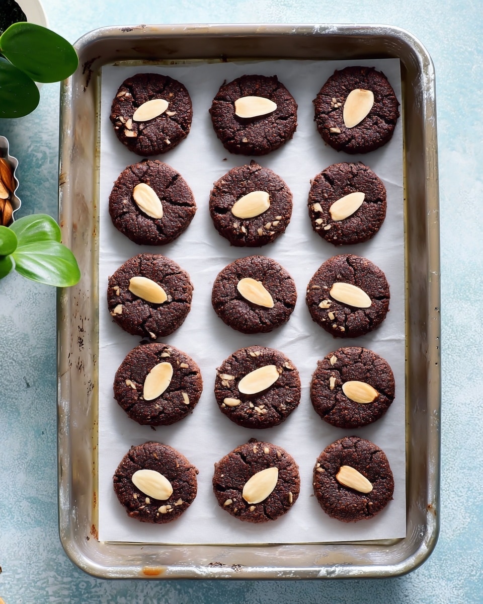 This image shows a metal baking tray lined with white parchment paper, holding twelve round chocolate cookies arranged in a 3 by 4 grid. Each cookie is dark brown with a rough texture and has a single pale almond slice placed flat in the middle. The tray rests on a light blue surface with a subtle white marbled texture. In the corners, small parts of other dishes and green leaves can be seen, but the focus remains on the tray of cookies photo taken with an iphone --ar 4:5 --v 7