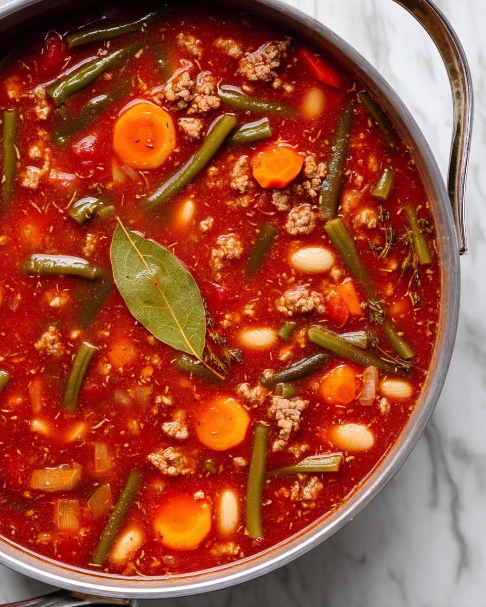 A large silver pot filled with a thick tomato-based soup, showing a rich red color with visible chunks of cooked ground meat, thin round orange carrot slices, green beans, and white beans scattered throughout. There are also pieces of onion and a bay leaf floating on top, adding texture and depth. The soup looks hearty with a mix of chunky and fine textures in a smooth red broth, all sitting on a white marbled surface photo taken with an iphone --ar 4:5 --v 7