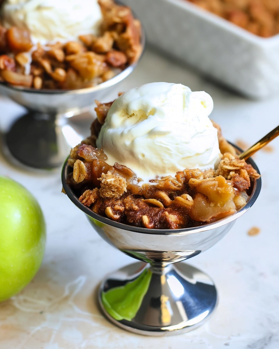 A close-up of a dessert served in a silver metal cup with a short stem, placed on a white marbled surface. The dessert has two layers: the bottom layer is a chunky apple crumble with golden brown baked pieces and visible bits of oats and nuts, with a slightly sticky and shiny texture, while the top layer is a smooth, round scoop of creamy white vanilla ice cream. Next to the cup is a gold spoon resting inside, and a green apple is partly visible to the left. The background includes a white textured baking dish with more apple crumble inside. Photo taken with an iphone --ar 4:5 --v 7