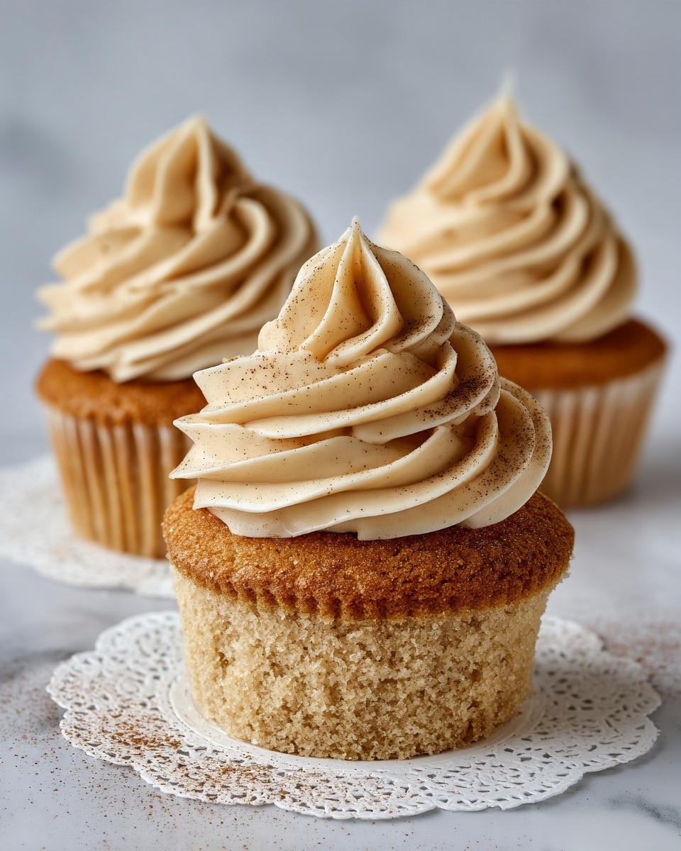 The image shows three cupcakes on a white doily placed on a white marbled surface. Each cupcake has one layer of light brown sponge cake with a slightly rough texture, topped with a swirl of pale tan frosting that is smooth and creamy. The frosting is piped in a spiral shape, standing tall above each cupcake. There is a light dusting of brown powder on the frosting and doily around the cupcakes. The photo is taken with an iphone --ar 4:5 --v 7