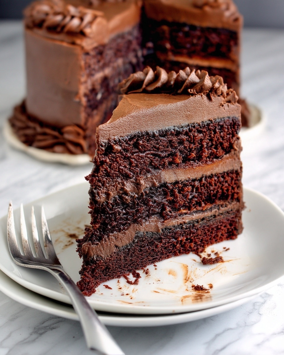 A slice of chocolate cake with three thick, dark brown layers of moist chocolate sponge separated by two layers of smooth, lighter brown chocolate frosting. The outside of the slice is covered with the same frosting, swirled and slightly textured, especially on the left edge. The slice rests on a white plate with traces of frosting and crumbs. A metal fork is partially visible on the left side of the plate. The background shows a blurred larger cake with the same chocolate layers. The surface beneath the plate has a white marbled texture. Photo taken with an iphone --ar 4:5 --v 7